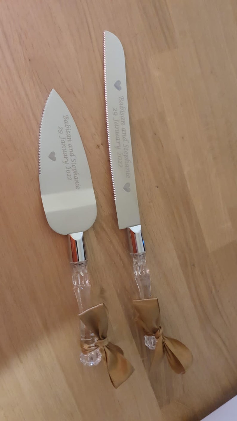 May include: A set of two silver cake knives with gold ribbon bows. The knives are engraved with the text "Babisan and Stephanie 29 January 2022".