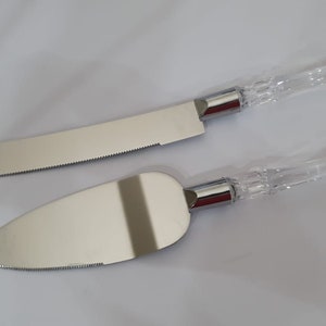 May include: A set of two silver cake knives with clear, crystal-like handles. The knives have serrated edges and are designed for cutting cake.