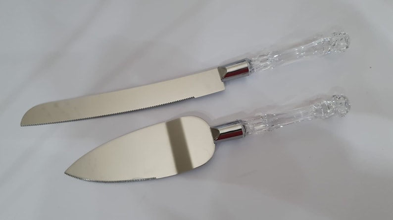 May include: A set of two silver cake knives with clear, crystal-like handles. The knives have serrated edges and are designed for cutting cake.