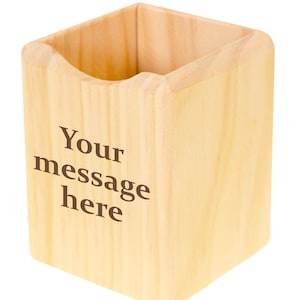 May include: A square wooden pen holder with a light natural grain finish. The front face features the dark brown printed text "Your message here" in a serif font. The container has an open top and rounded vertical edges on the front side.