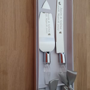 May include: A set of two silver cake knives with crystal handles and gray satin bows. The knives are engraved with "Jason & Selena 10th March 2022".