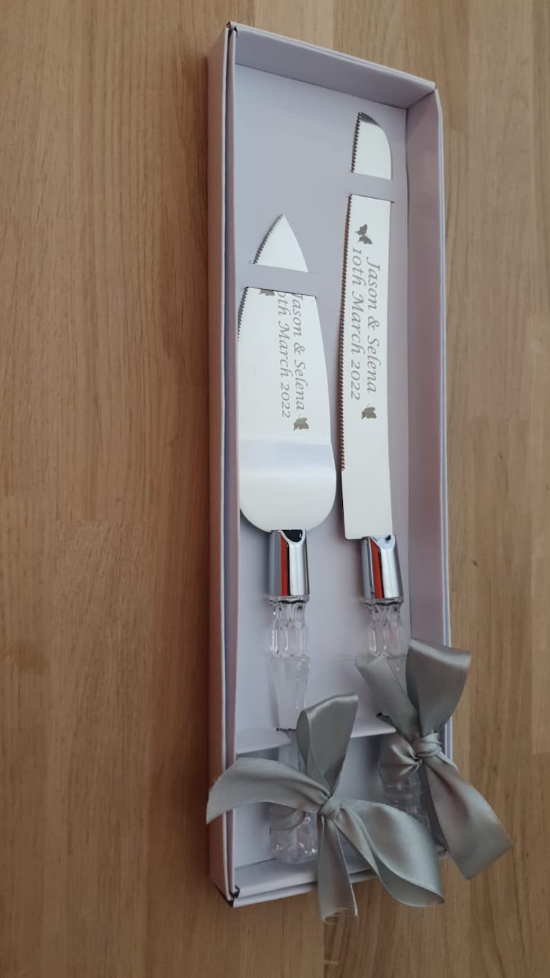 May include: A set of two silver cake knives with crystal handles and gray satin bows. The knives are engraved with "Jason & Selena 10th March 2022".