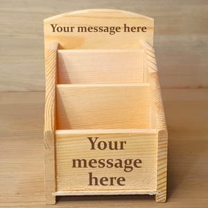 May include: A light-coloured wooden desk organiser with three tiered compartments for holding items. The top back panel and the front panel both feature the dark brown engraved text "Your message here" in a clear, serif font style.