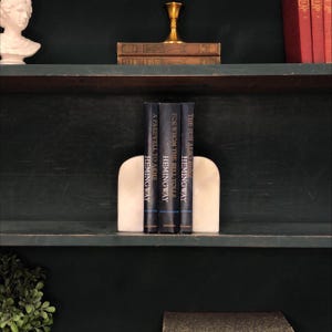 May include: A shelf displays three books by Hemingway, held by white marble bookends. The book titles are "A Farewell to Arms", "For Whom the Bell Tolls", and "The Sun Also Rises". The books are dark blue with white text. A bust and other books are visible.