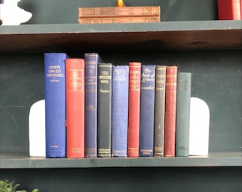 May include: A collection of antique books in various colors, including blue, red, and green, arranged on a shelf. The books are held in place by white bookends. A small gold-colored decorative object sits on the shelf above.
