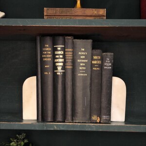 Pu&ograve; includere: Una collezione di libri d'epoca con copertine nere e scritte dorate, tenuti tra due fermalibri in marmo bianco. I titoli includono "The Search for the Ancient Order" e "The People's New Testament".
