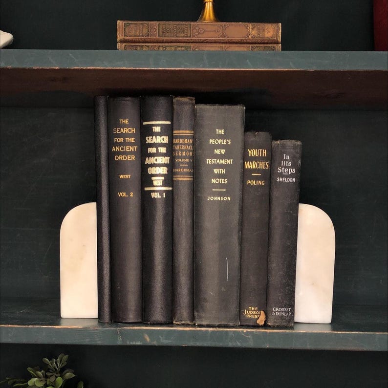 Pu&ograve; includere: Una collezione di libri d'epoca con copertine nere e scritte dorate, tenuti tra due fermalibri in marmo bianco. I titoli includono "The Search for the Ancient Order" e "The People's New Testament".