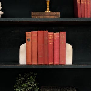 May include: A collection of seven vintage books with red and orange cloth covers, held between two smooth white marble bookends. The spines feature gold lettering with titles including The Inner Shrine, Hearts Courageous, and A Daughter of the South.