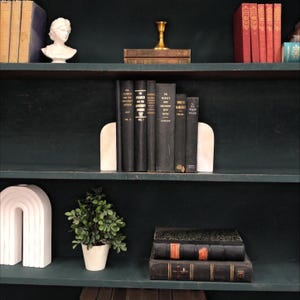 Pu&ograve; includere: Una libreria verde scuro espone una collezione di libri d'epoca, fermalibri in marmo e oggetti decorativi. I libri sono rilegati in nero e rosso, con dettagli dorati. Una piccola pianta in vaso e un busto bianco completano l'estetica.