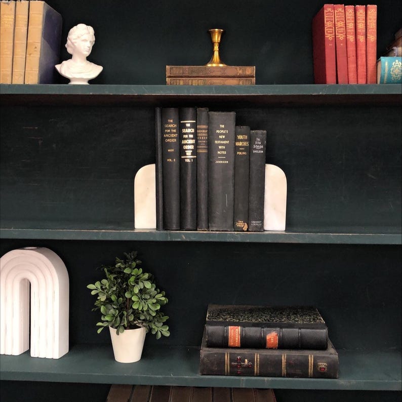 Pu&ograve; includere: Una libreria verde scuro espone una collezione di libri d'epoca, fermalibri in marmo e oggetti decorativi. I libri sono rilegati in nero e rosso, con dettagli dorati. Una piccola pianta in vaso e un busto bianco completano l'estetica.