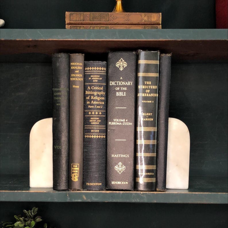 Może przedstawiać: Kolekcja starych książek z ciemnymi okładkami, w tym słownik i tomy o religii i teologii, jest trzymana między dwoma białymi marmurowymi podp&oacute;rkami do książek na p&oacute;łce.