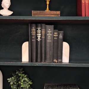 Pu&ograve; includere: Una mensola scura mostra una collezione di libri scuri, tenuti in posizione da fermalibri in marmo bianco. I libri hanno scritte dorate e titoli come "The Search for the Ancient Order" e "Youth Marches".