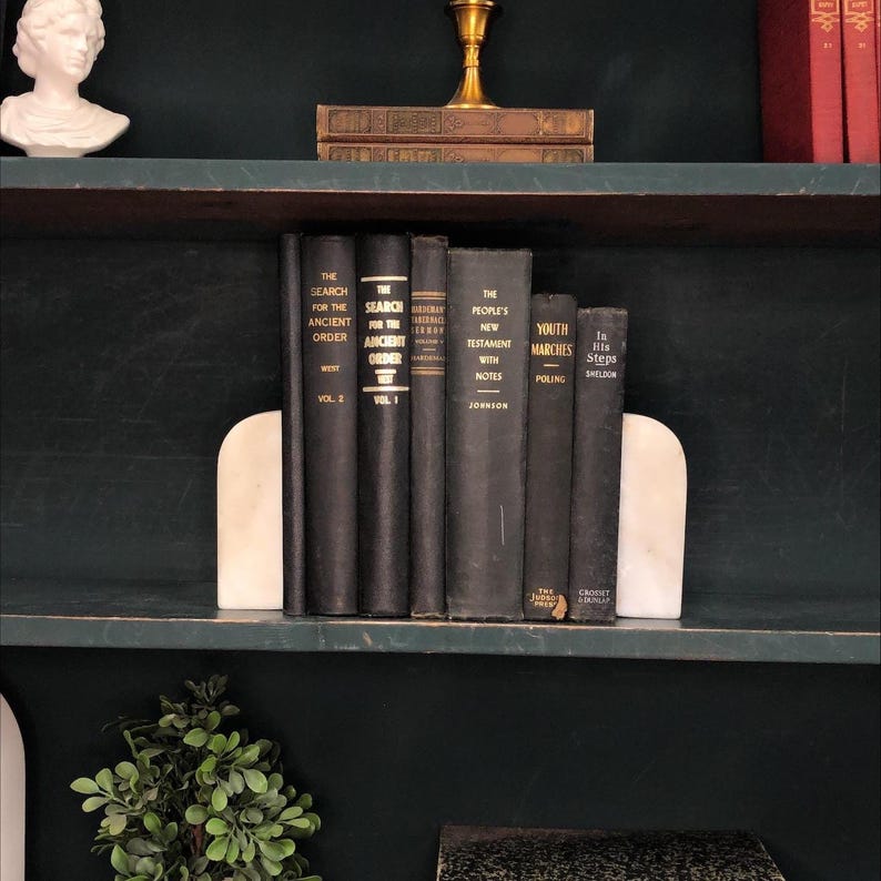 Pu&ograve; includere: Una mensola scura mostra una collezione di libri scuri, tenuti in posizione da fermalibri in marmo bianco. I libri hanno scritte dorate e titoli come "The Search for the Ancient Order" e "Youth Marches".