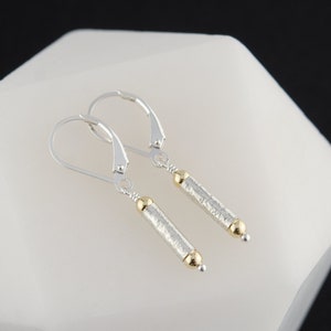 以下が含まれることがあります： ゴールドのアクセントが付いたシルバーのドロップイヤリングのペア。イヤリングは、両端に小さなゴールドビーズが付いた、テクスチャードのシリンダー形状をしています。