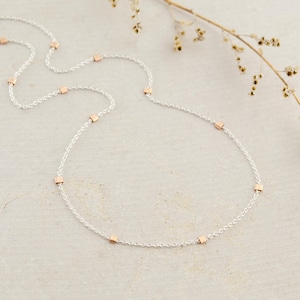May include: A fine silver necklace with evenly spaced rose gold square beads. The necklace is shown on a neutral surface, with a sprig of dried flowers in the background. The chain is delicate and elegant, suitable for everyday wear.