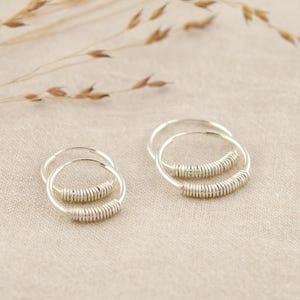 May include: Two pairs of silver hoop earrings. Each earring has a coiled design in the centre. The earrings are displayed on a neutral fabric, with dried grass in the background.