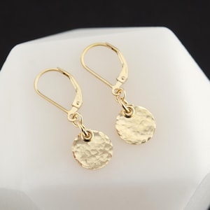 May include: A pair of gold-coloured leverback earrings. Each earring has a hammered, circular charm suspended from a leverback closure. The earrings are displayed on a white surface with a dark background.