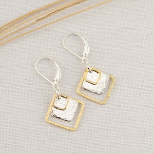 May include: A pair of silver and gold earrings with a geometric design. Each earring features a silver hammered square within a gold square frame. The earrings have a lever back closure and are displayed on a white surface.