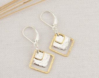 May include: A pair of silver and gold earrings with a geometric design. Each earring features a silver hammered square within a gold square frame. The earrings have a lever back closure and are displayed on a white surface.