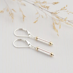 May include: A pair of silver dangle earrings with gold accents. The earrings feature a textured, cylindrical charm.