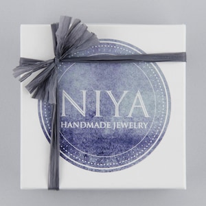 Puede incluir: Una caja de regalo cuadrada de color blanco atada con una cinta de rafia gris oscuro. Centrada en la caja hay una etiqueta circular de estilo acuarela azul con el texto blanco NIYA HANDMADE JEWELRY en fuente serif, rodeada por un borde punteado.