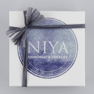 以下が含まれることがあります： グレーのリボンと弓が付いた白いギフトボックス。ボックスには、"NIYA HANDMADE JEWELRY"というテキストが印刷された青と白の水彩円があります。