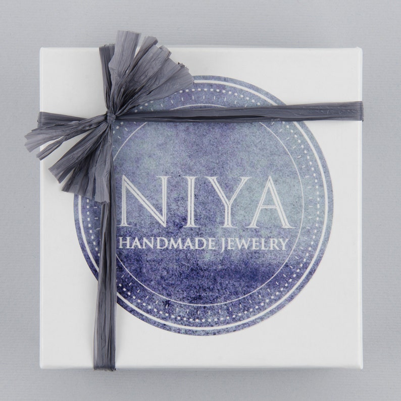以下が含まれることがあります： グレーのリボンと弓が付いた白いギフトボックス。ボックスには、"NIYA HANDMADE JEWELRY"というテキストが印刷された青と白の水彩円があります。