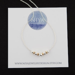 Puede incluir: Un delicado collar de cadena plateada con una disposici&oacute;n central de peque&ntilde;as cuentas met&aacute;licas que alternan entre plateado y dorado. El collar se muestra sobre una tarjeta blanca con el texto NIYA HANDMADE JEWELRY y WWW.NIYAJEWELRYDESIGN.ETSY.COM.