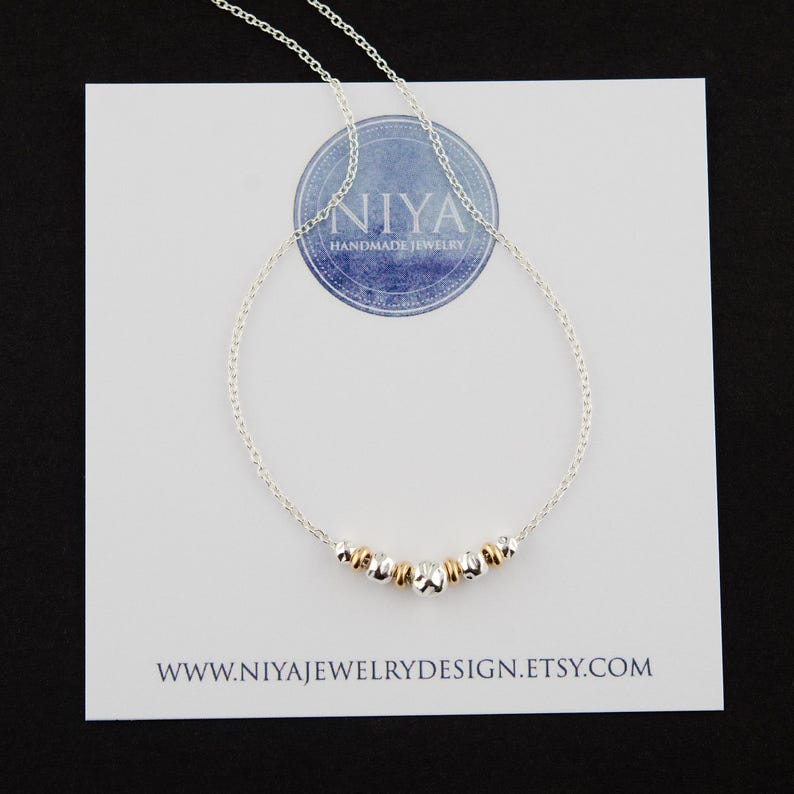 Puede incluir: Un delicado collar de cadena plateada con una disposici&oacute;n central de peque&ntilde;as cuentas met&aacute;licas que alternan entre plateado y dorado. El collar se muestra sobre una tarjeta blanca con el texto NIYA HANDMADE JEWELRY y WWW.NIYAJEWELRYDESIGN.ETSY.COM.