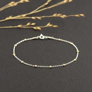 May include: A delicate bracelet featuring a thin silver chain interspersed with small, evenly spaced gold-toned cube beads. The bracelet is secured with a circular spring ring clasp, creating a refined and balanced design with metallic contrast.