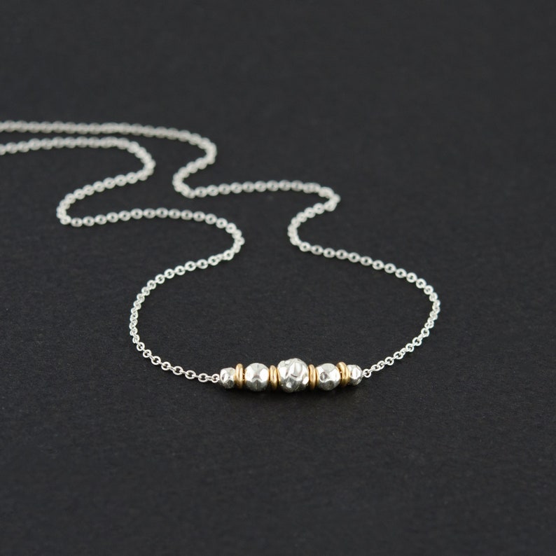 May include: A delicate silver chain necklace featuring a central focal point composed of three textured silver beads interspersed with two small gold-toned spacer beads, creating a subtle contrast between the metallic finishes and the varied bead surfaces.