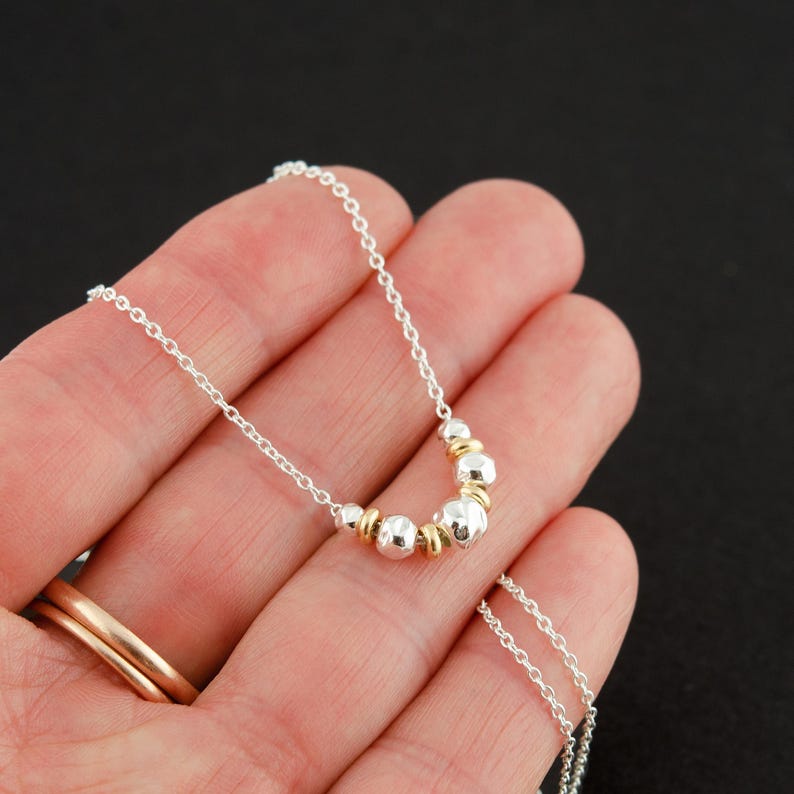 Puede incluir: Un collar de cadena de plata delicada con una disposici&oacute;n central de peque&ntilde;as cuentas de plata martillada que se alternan con anillos espaciadores lisos y pulidos de tono dorado. El collar tiene una construcci&oacute;n de eslabones finos.