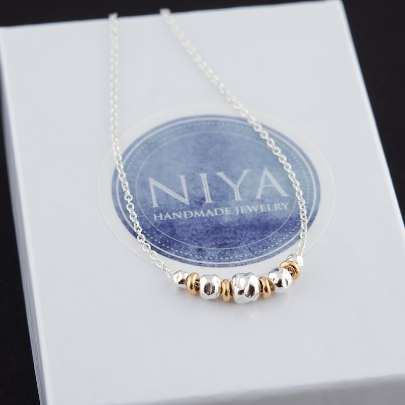 Puede incluir: Un delicado collar de cadena plateada con una disposici&oacute;n central de peque&ntilde;as cuentas plateadas texturizadas que alternan con espaciadores de anillo lisos de tono dorado. El collar se muestra sobre una caja blanca con un logotipo circular azul que dice NIYA.