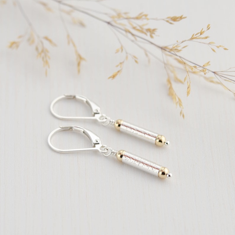 May include: A pair of silver dangle earrings with gold accents. The earrings feature a textured, cylindrical charm.