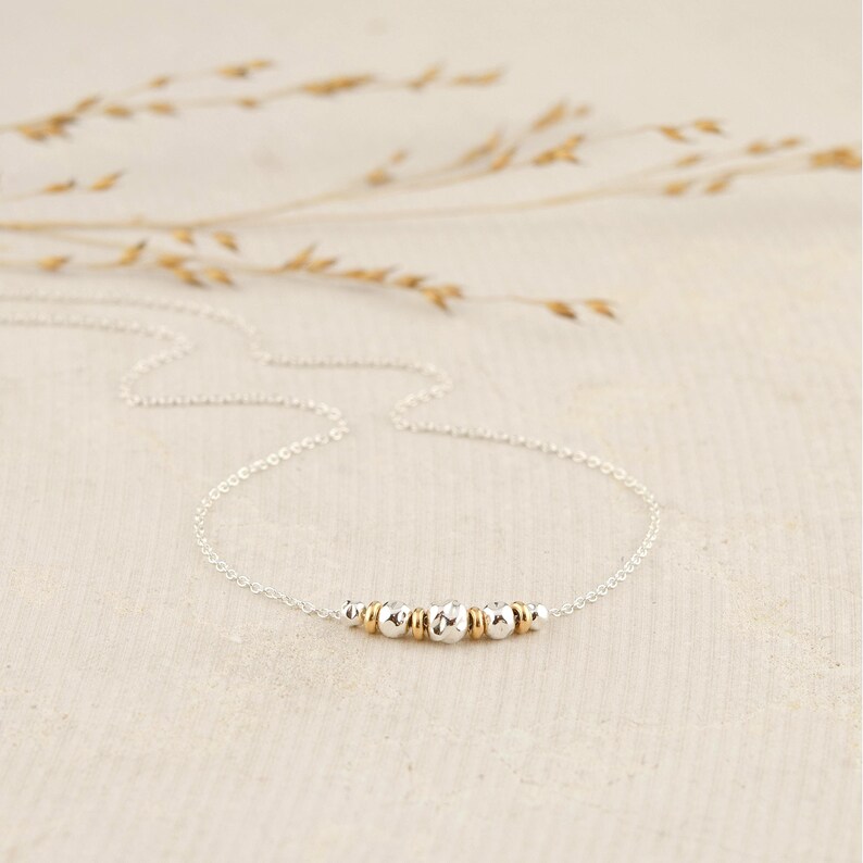 Puede incluir: Un collar de cadena plateada fina que presenta una disposici&oacute;n central de peque&ntilde;os anillos dorados y cuentas plateadas texturizadas alternadas. El collar tiene un dise&ntilde;o de cadena de eslabones finos que conecta con el grupo central de cuentas.