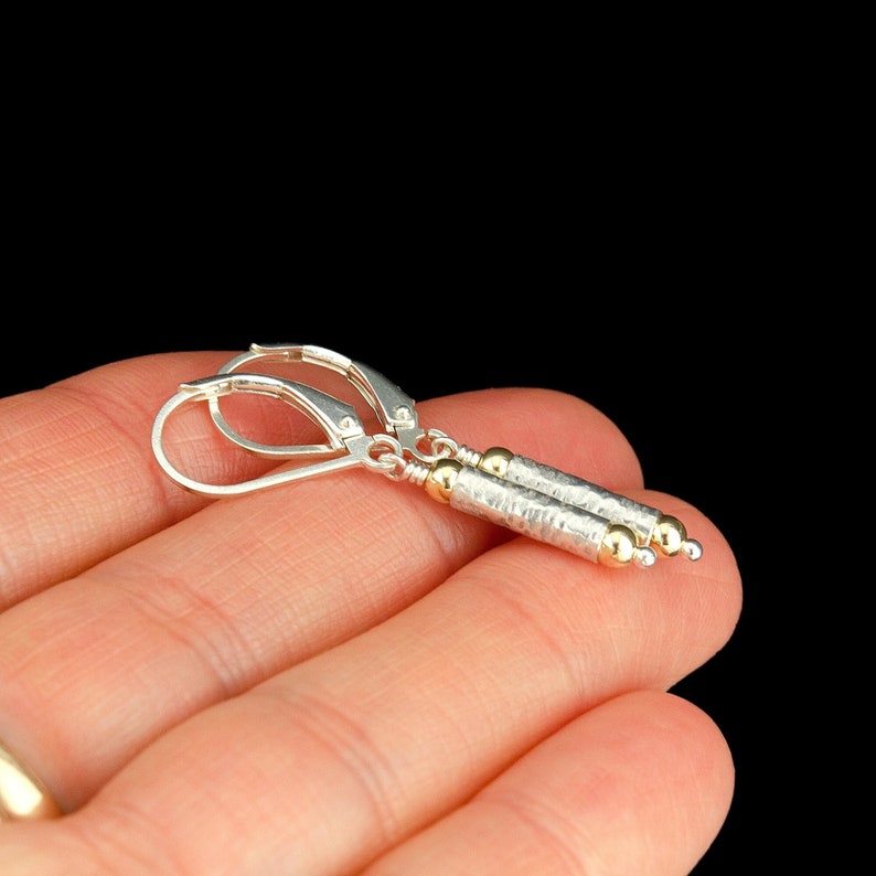 以下が含まれることがあります： ゴールドのアクセントが付いたシルバーのドロップイヤリング。イヤリングは、ゴールドビーズがついたテクスチャードシルバーのシリンダーが2つ付いています。イヤリングは、シルバーのレバーバック留め具に付いています。