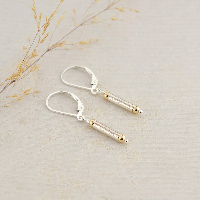 May include: A pair of silver earrings with gold accents. Each earring features a textured, cylindrical pendant with a small gold bead at the end.