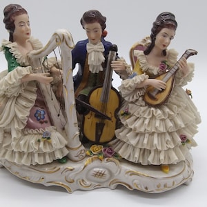 May include: A porcelain figurine set of three people playing musical instruments. A woman plays a harp, a man plays a cello, and a woman plays a mandolin. The figurines are dressed in elaborate clothing and are set on a white base with gold accents.