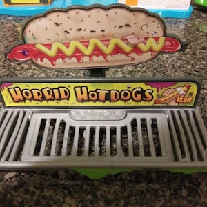 May include: A toy hot dog stand with a green and yellow sign that reads "HORRID HOTDOGS" and a price of £2.50. The stand has a grey metal grill and a hot dog with mustard and ketchup on top.