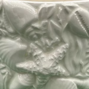 Puede incluir: Maceta de cerámica blanca con un diseño en relieve de conchas y flores. La maceta es de un tono blanco claro y tiene un acabado liso y brillante.