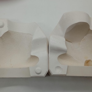 Pode incluir: Dois moldes de cerâmica branca para fundir um objeto tridimensional. Os moldes estão abertos e mostram o detalhe interior do objeto.