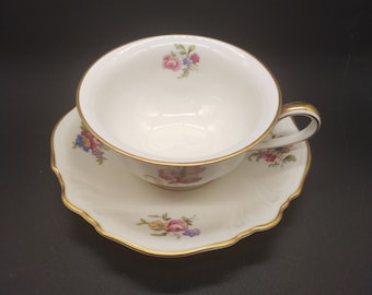 May include: A white teacup and saucer set with a floral pattern in shades of pink, blue, and yellow. The set has a gold trim around the edges.