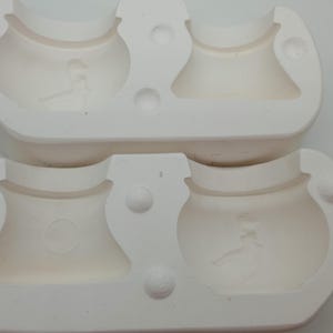 May include: A white ceramic mould for making two small cauldron-shaped objects. The mould is in two pieces, and the top piece has two indentations for the cauldrons. The bottom piece has two indentations for the cauldrons, and the two pieces fit together to create a complete mould.