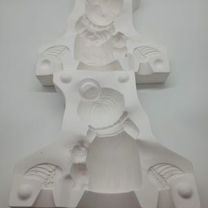 May include: Two white ceramic molds, each featuring a detailed relief of a child holding a teddy bear. The molds have a decorative shape with wing-like details on the sides. The overall design is delicate and suitable for crafting.