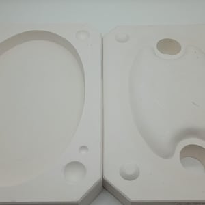 May include: Two white plaster molds, one oval and one teardrop-shaped, are shown side-by-side. Each mold has a raised border and several small, round indentations. The molds are likely used for casting or creating objects.
