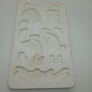 May include: A white ceramic mould with a design of two swans, a bow, and other small shapes. The mould is labelled "No. 201 - Colorama - Switzerland - Art No. 1000 - Made in Switzerland - 1986".