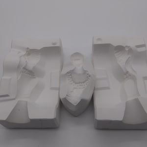 May include: Three white plaster molds for creating decorative sculptures. The molds depict a woman in a dress and a bust of a woman with a necklace. The molds are arranged on a white surface.
