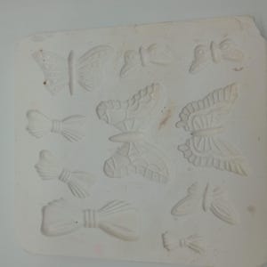 Peut inclure: Un moule carré blanc avec divers motifs de papillons et de nœuds papillon. Le moule présente plusieurs formes de papillons avec des motifs d'ailes détaillés et des formes de nœuds papillon. Les motifs sont en relief, créant un effet 3D. Le moule est probablement destiné à l'artisanat ou à la pâtisserie.