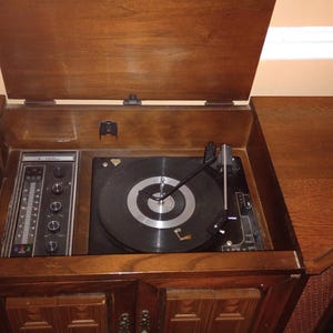 May include: A wooden console record player with a hinged top lid, featuring a black turntable, a black tonearm, and a vertical control panel with four circular knobs and a linear radio frequency display scale set within a dark brown cabinet.