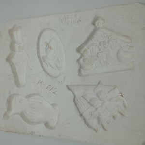 Peut inclure: Moule en céramique blanche avec cinq motifs différents sur le thème des fêtes. Les motifs comprennent un bonhomme de neige, un pingouin, un sapin de Noël, une maison et une figurine tenant une cloche. Le moule est étiqueté "Mock's Notions" et "M-612".